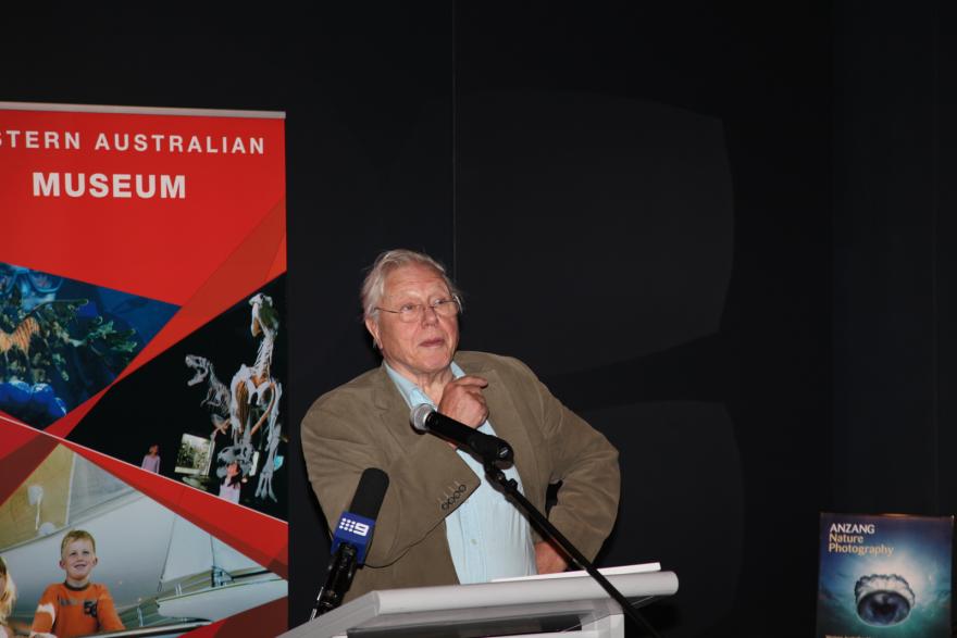Person speaking at a podium with a red banner and a nature photography poster in the background.