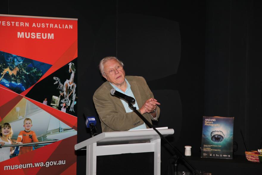 Person speaking at a podium with a red banner and a nature photography poster in the background.