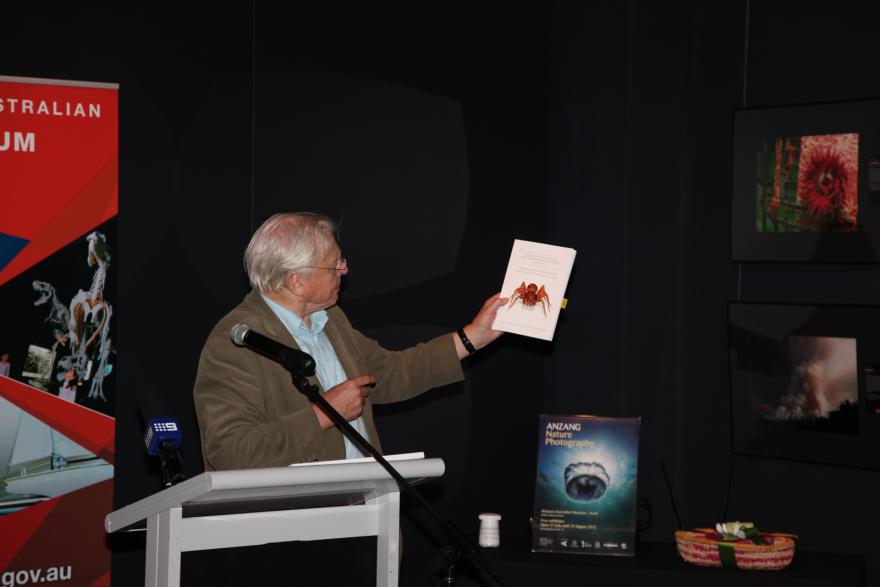 Person speaking at a podium with a red banner and a nature photography poster in the background, whilst holding up a picture of a spider..