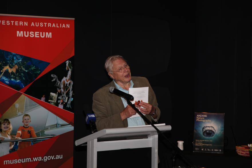 Person speaking at a podium with a red banner and a nature photography poster in the background, whilst holding up a picture of a spider..