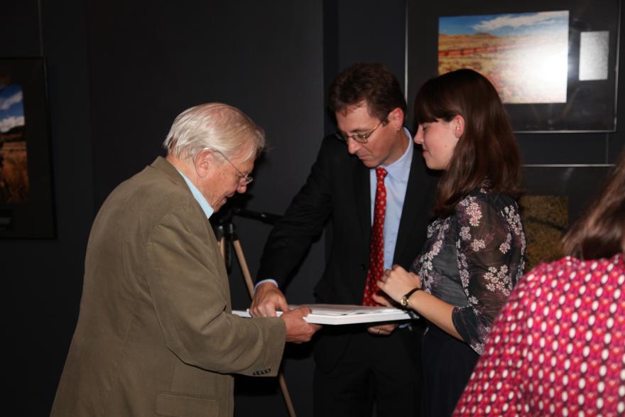 Two people are talking in front of a dark background; one wearing a brown jacket and glasses, and the other wears a floral blouse. There is another person wearing a suit standing with them.