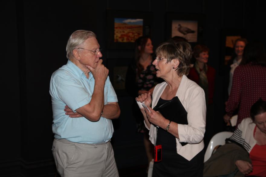 A person in a light blue shirt talks with another person holding a notebook in a dimly lit room.