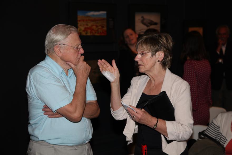 A person in a light blue shirt talks with another person holding a notebook in a dimly lit room.