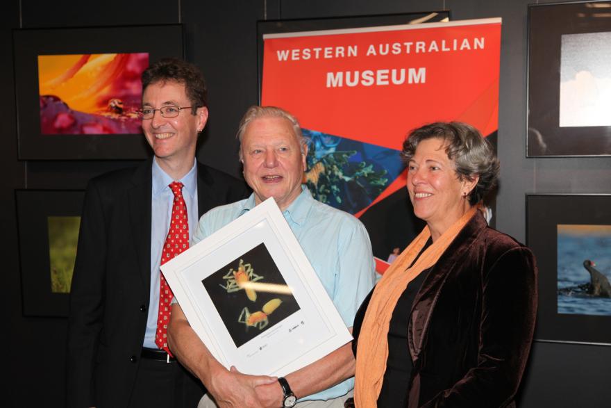 Three people posing with a framed photograph in front of a "Western Australian Museum" backdrop.