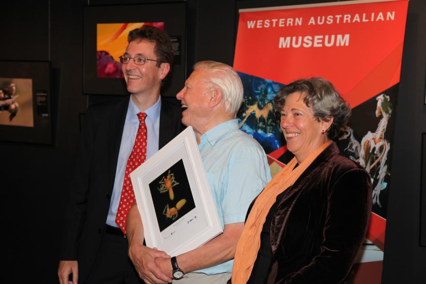 Three people posing with a framed photograph in front of a "Western Australian Museum" backdrop.