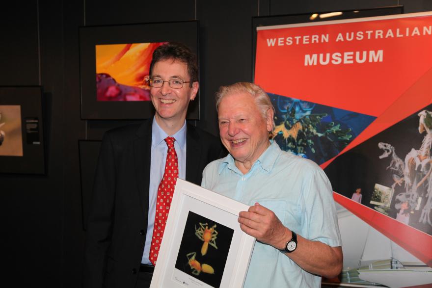 Two smiling people pose with a framed photograph in front of a Western Australian Museum display.
