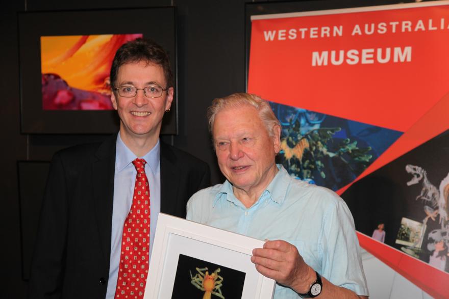 Two smiling people pose with a framed photograph in front of a Western Australian Museum display.
