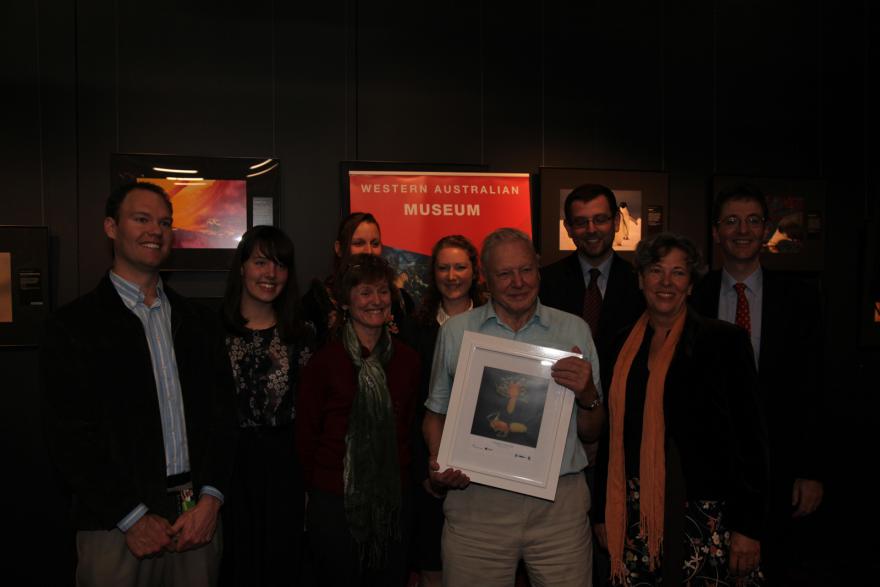 A group of nine people posing in front of a "Western Australian Museum" sign, with one person holding a framed picture.