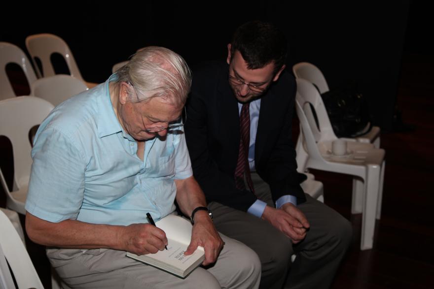 Two people, one signing a book, seated in white chairs.