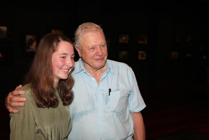 Two people smiling together indoors, with framed pictures in the dark background.