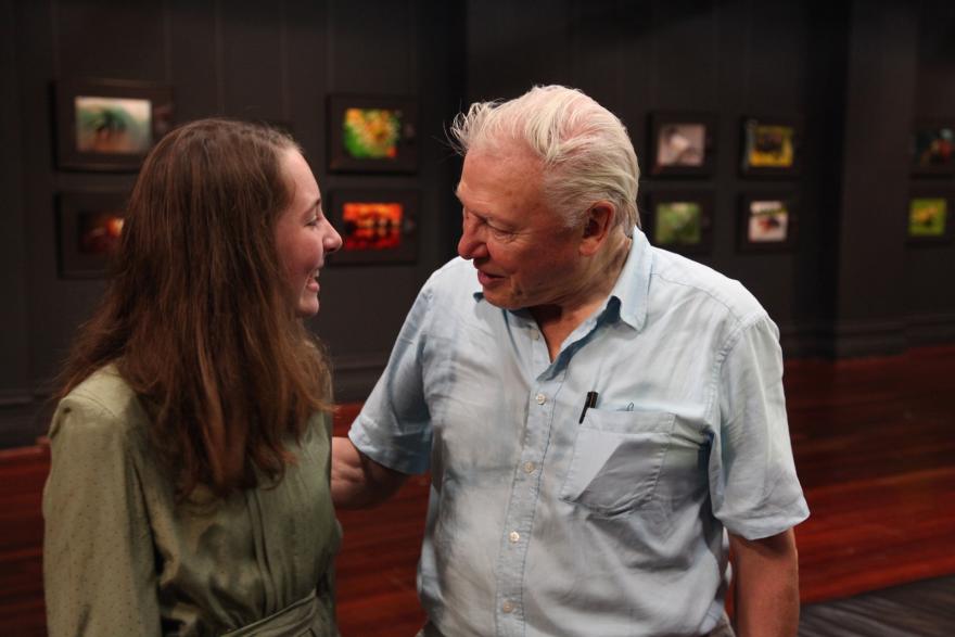 Two people smiling together indoors, with framed pictures in the dark background.