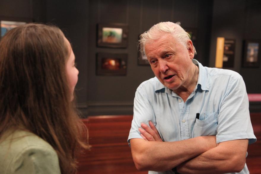 Two people talking together indoors, with framed pictures in the dark background.