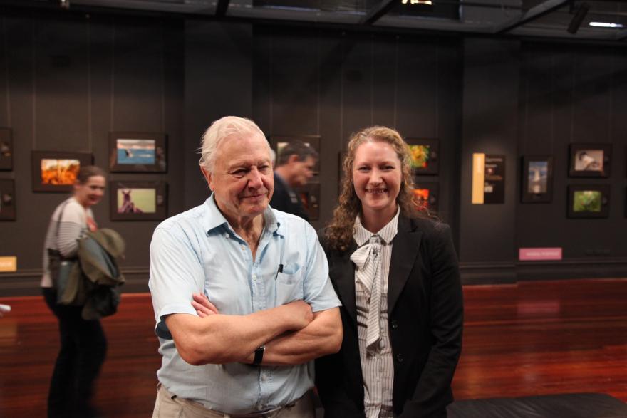 Two people smiling in a gallery setting with framed pictures on the walls behind them.