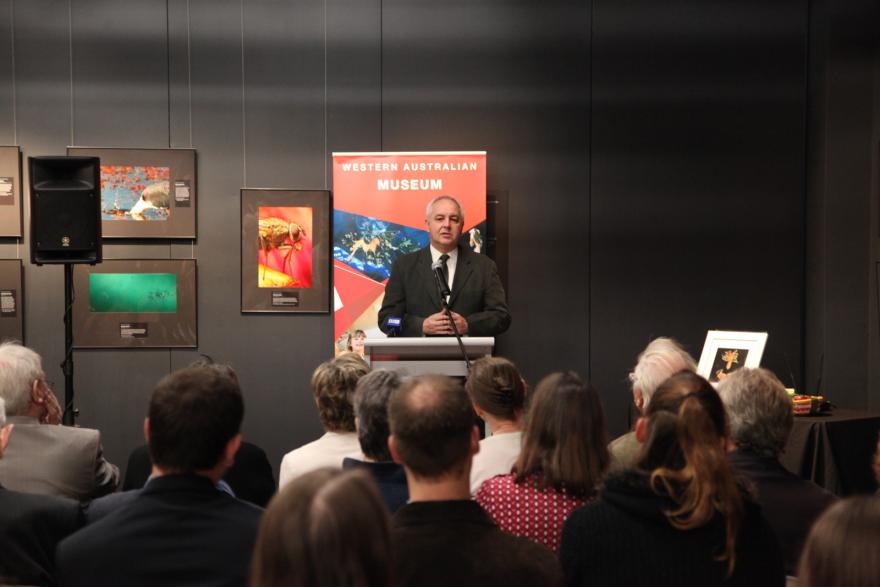 A person speaking at a podium in front of a "Western Australian Museum" banner, with photographs on display behind him and an audience seated.