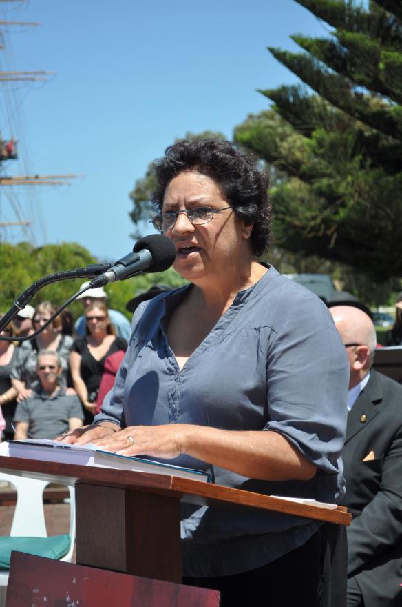 A person in a blue shirt speaks at an outdoor event behind a podium with a microphone.