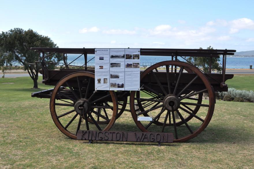 A vintage wagon with a display board, labeled "KINGSTON WAGON," set against a scenic backdrop near water.