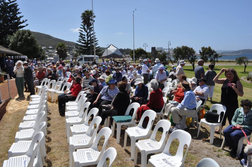 Outdoor gathering with seated people on white chairs, some marked "Reserved," against a scenic backdrop with trees and water.