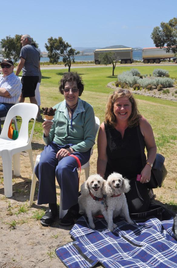 Two people with small white dogs sitting outdoors by the water.