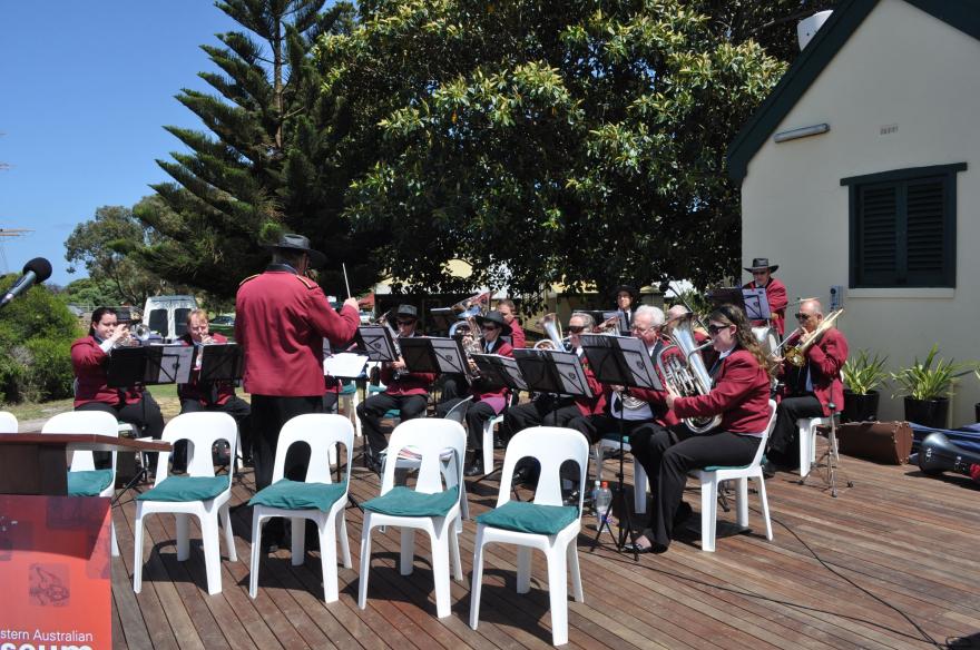 Outdoor band performance with musicians in maroon jackets playing brass instruments, a conductor leading in the center, surrounded by greenery.