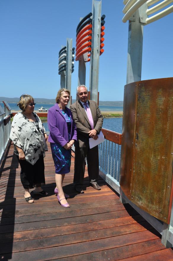Three people standing on a boardwalk beside a metal structure with colourful rods near water.