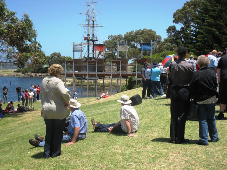 People gathered on a grassy area near a body of water, with a ship mast structure in the background.