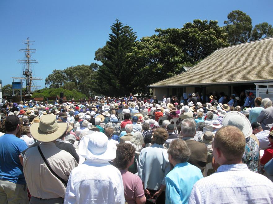 A large crowd wearing hats gathered outside a building with a ship's mast and trees in the background under a clear blue sky.