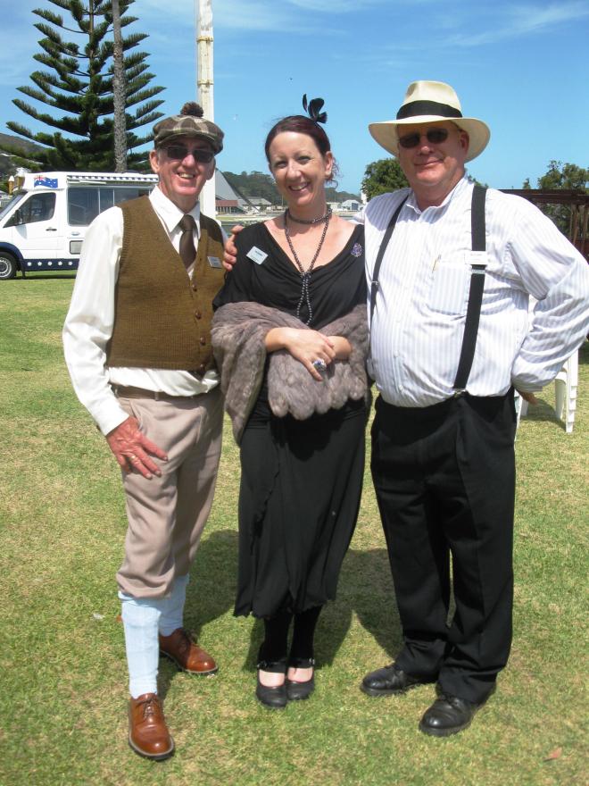 Three people dressed in vintage clothing standing on grass with a clear sky and a vehicle in the background.