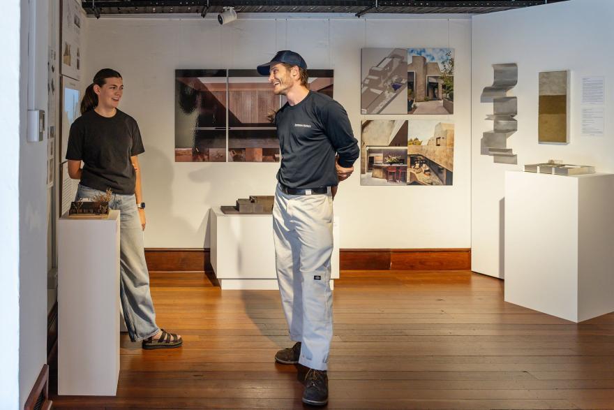 Two people in black shirts and light trousers smile at each other in a brightly lit gallery decorated with handing images and objects on white plinths