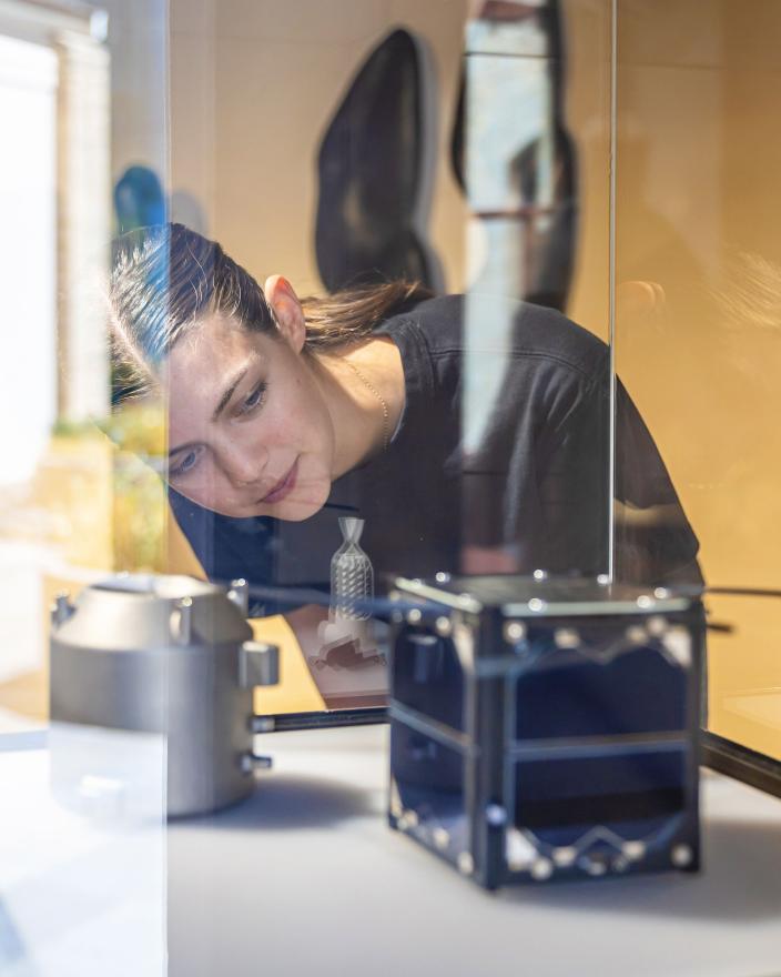 A person with brown hair and a black shirt looks at a small square object in a glass showcase