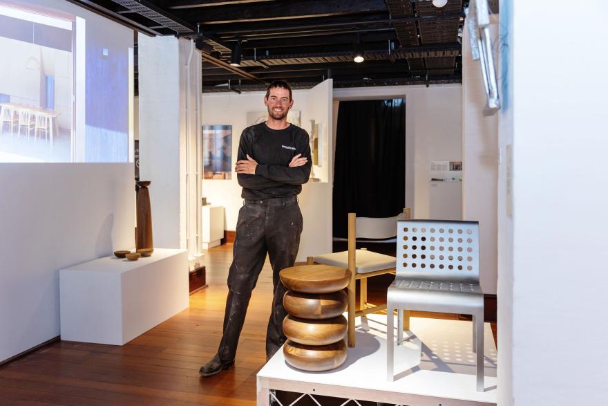 A smiling person wearing black trousers and a black shirt stands with their arms crossed in a brightly lit gallery featuring stylistic pieces of small furniture