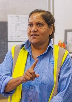 Person in a blue shirt and yellow safety vest gesturing indoors.