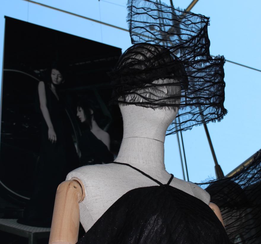 A mannequin in a black dress with sheer fabric on its head, set against a backdrop with a photograph of two figures.