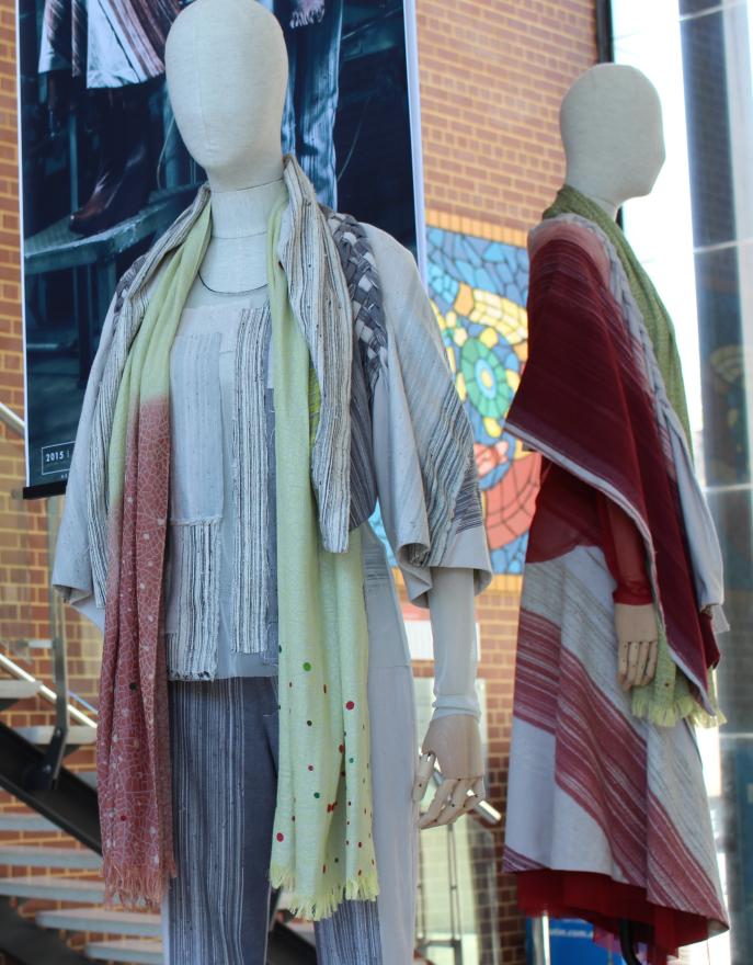 Two mannequins in layered, textured outfits with scarves and capes, standing in front of a brick wall and colorful mosaic.