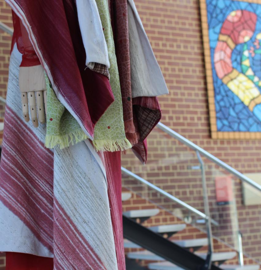Mannequin draped in colorful fabrics near a brick wall and staircase with a mosaic artwork in the background.