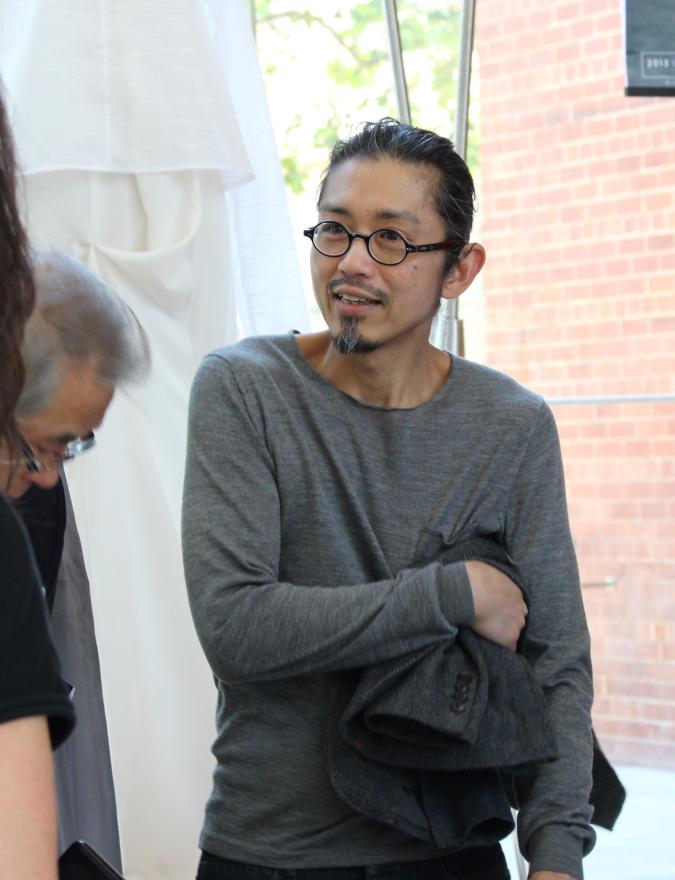 A person wearing glasses and a gray shirt holds a dark jacket, standing near a large window with brick and fabric background.