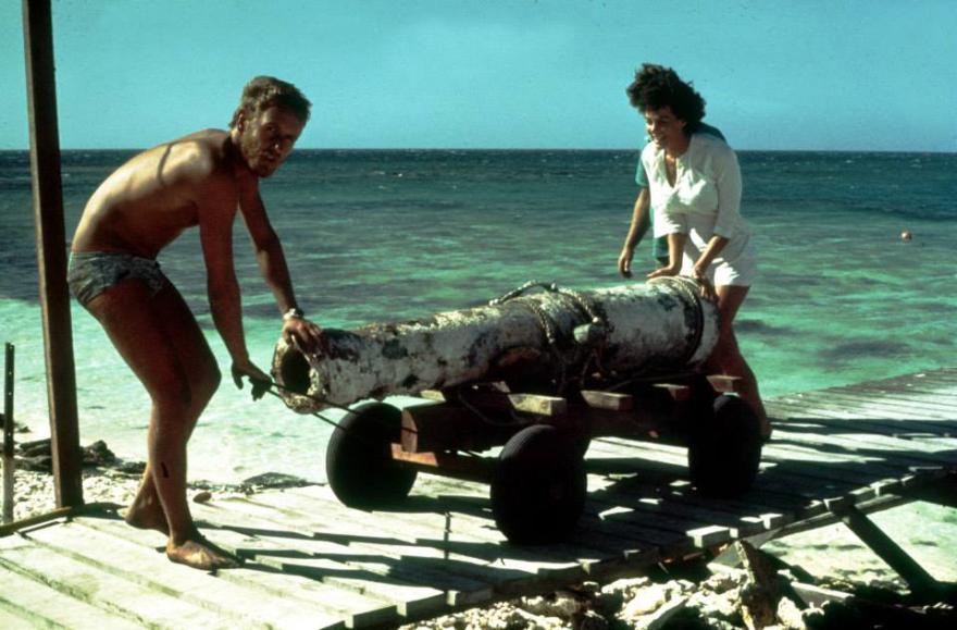Two people moving a large cannon on a wheeled cart by the ocean on a wooden dock.