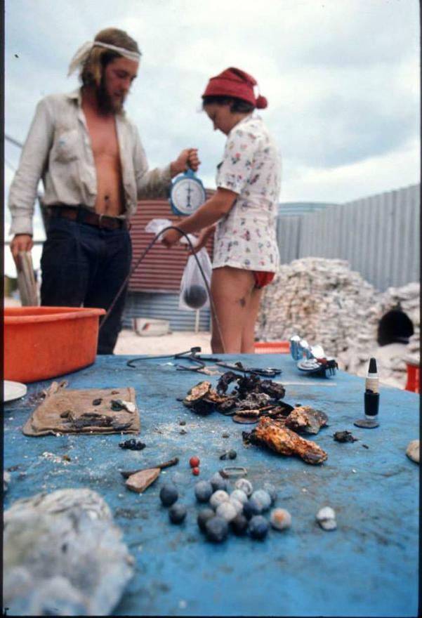 Two people outdoors by a blue table with various small objects, including shells and stones.