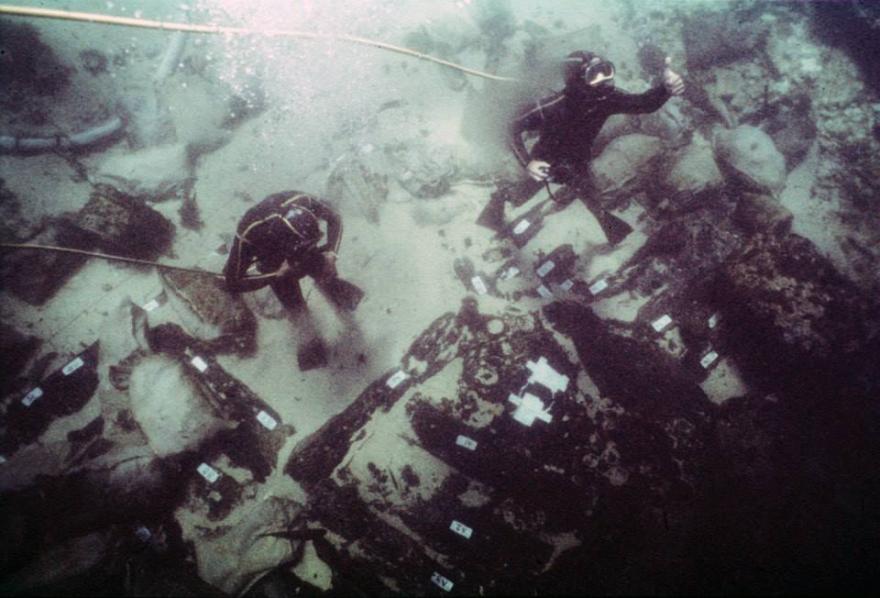 Two divers examining a shipwreck underwater, surrounded by sediment-covered artifacts with white labels.