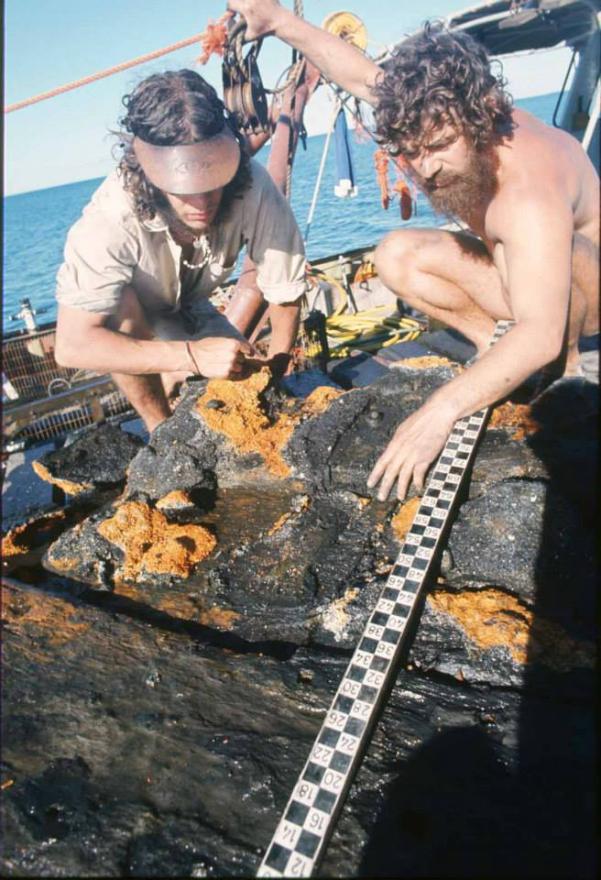 Two people examining a black object with orange patches on a boat deck.
