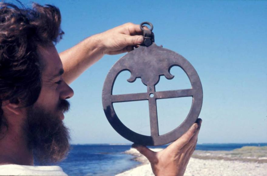 Person holding a circular astrolabe against a blue sky with a sea and shoreline in the background.