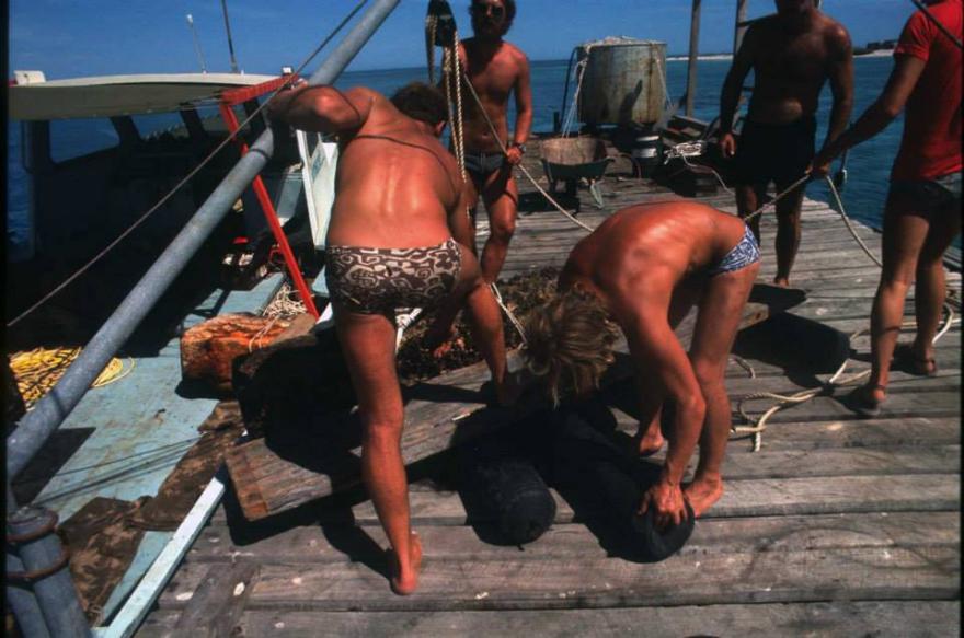 People on a dock lifting a large cylindrical object with a boat and sea in the background.
