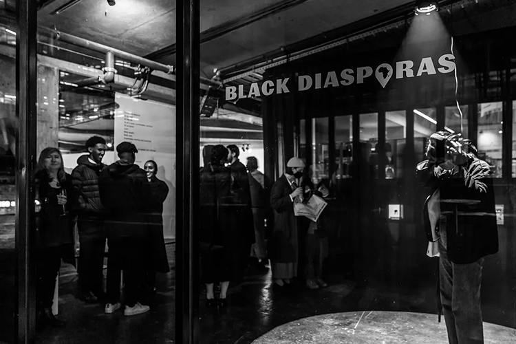 A black and white photo of a gallery with the text "BLACK DIASPORAS" on a glass wall, showing people inside engaged in conversation.