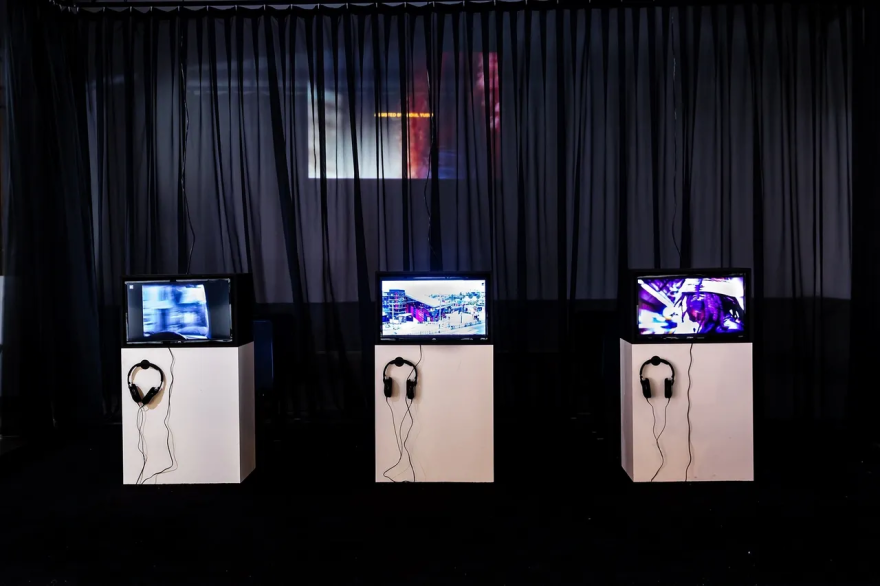 Three televisions on pedestals with headphones in a dimly lit exhibition room.