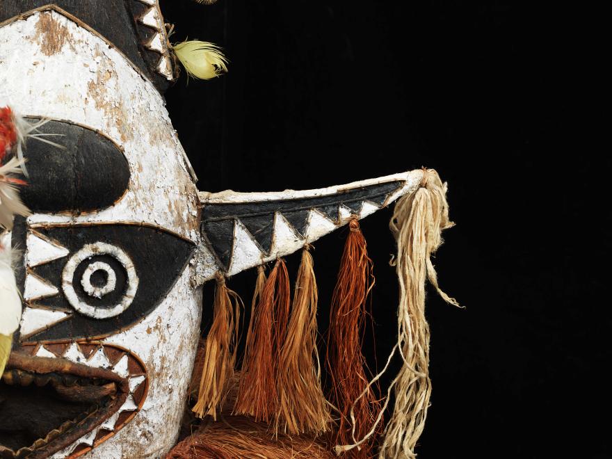 Close-up of a tribal mask with black, white, and beige patterns, featuring circular and triangular designs, tassels, and feathers.
