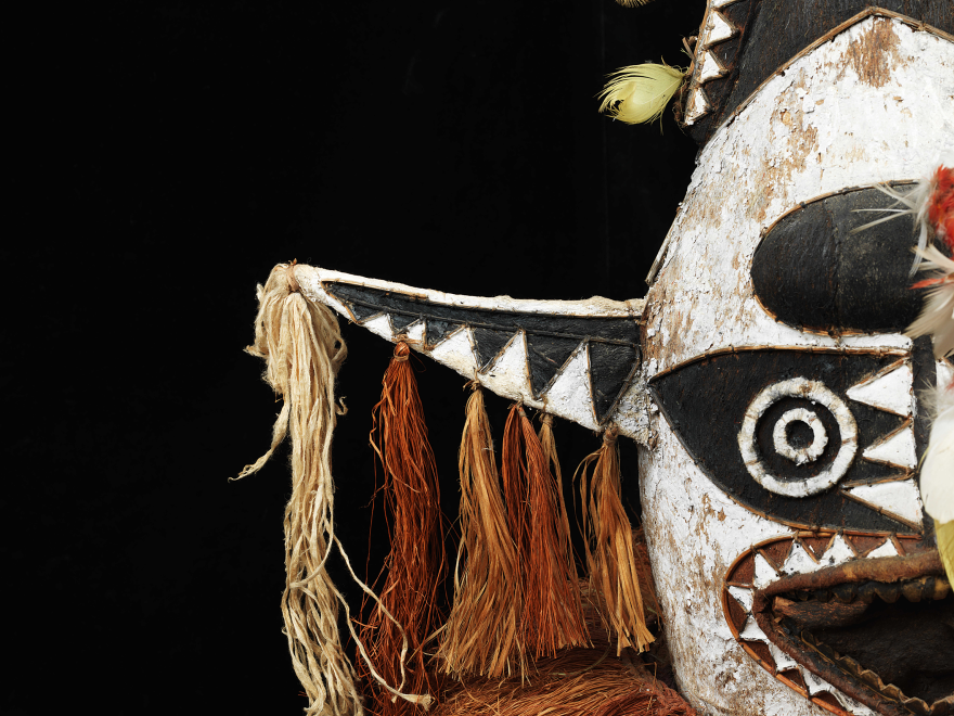 Close-up of a tribal mask with black, white, and beige patterns, featuring circular and triangular designs, tassels, and feathers.