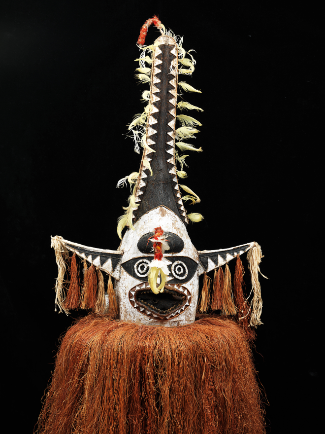 Close-up of a tribal mask with black, white, and beige patterns, featuring circular and triangular designs, tassels, and feathers.