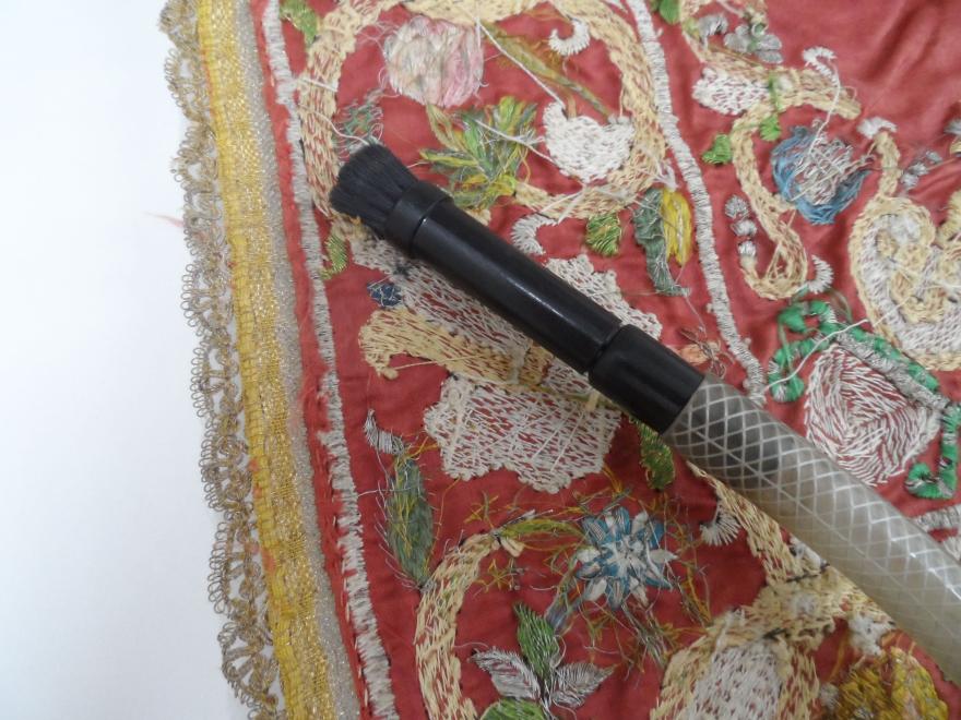 Intricately embroidered textile with a lace border and a dark-handled dust vacuum placed diagonally on it.