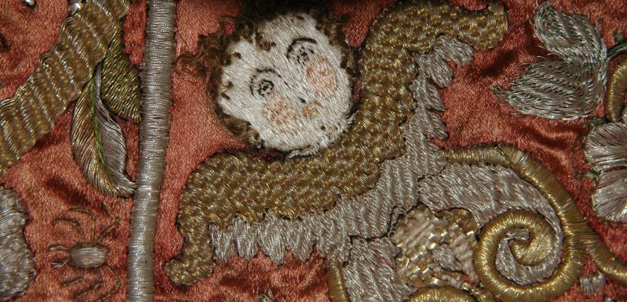 Close-up of an elaborate embroidery with a cherubic face and gold thread designs on a reddish fabric.
