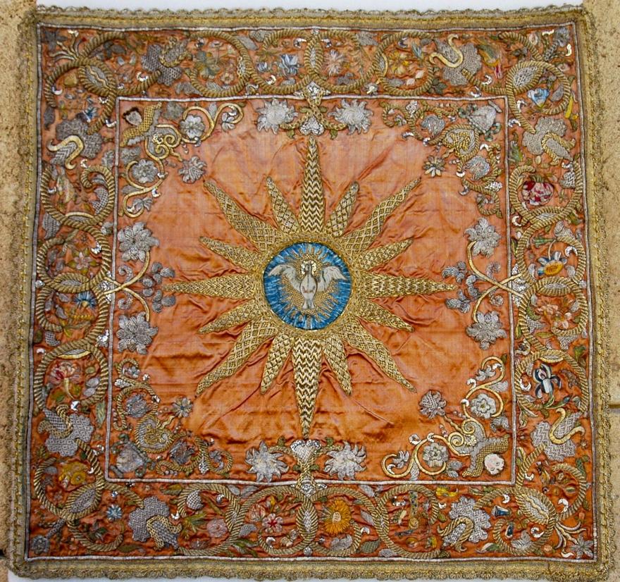 An embroidered textile with a central pelican emblem and sunburst on an orange background, surrounded by floral motifs.