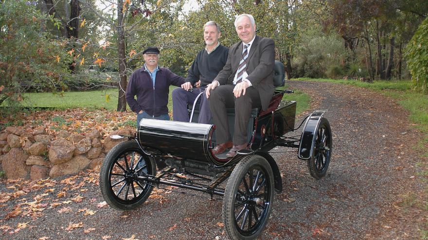 Three men with a vintage black automobile on a gravel path, surrounded by trees with autumn leaves.